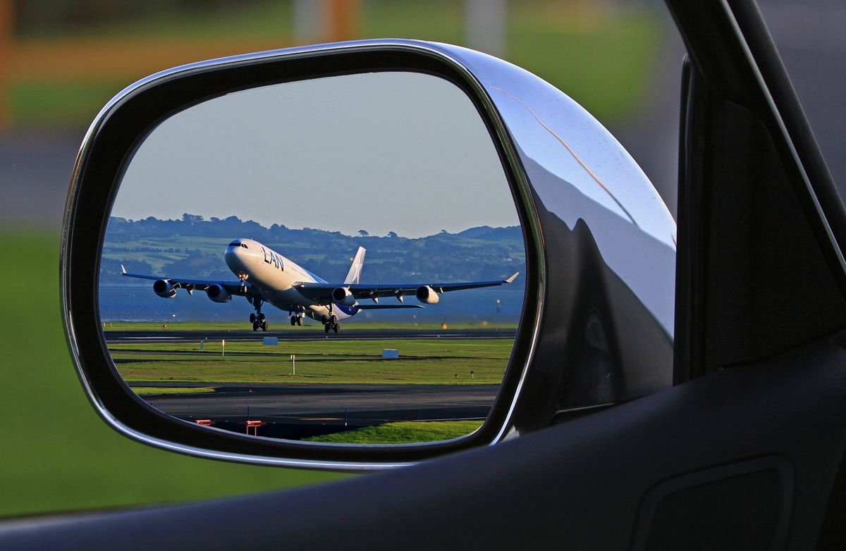 A jet taking off reflected in the side mirror of a car at an airport