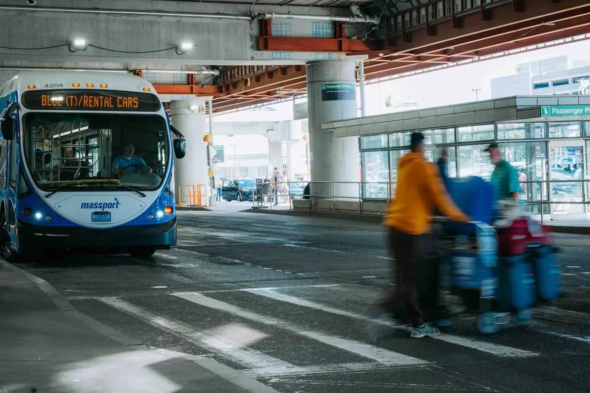 Travellers walking past a rental car shuttle bus waiting under an airport terminal canopy